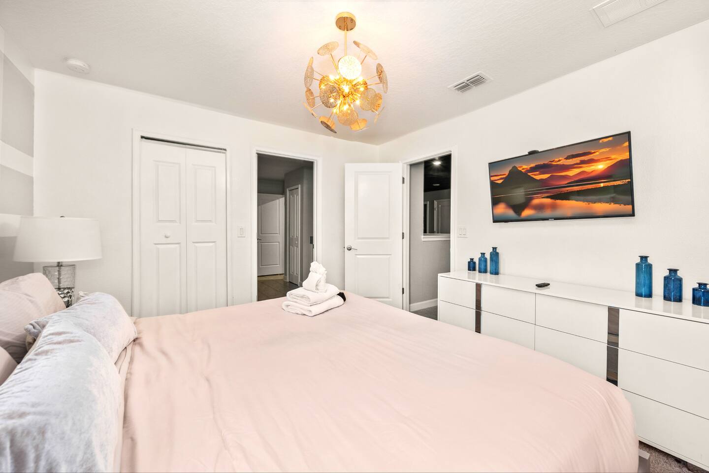 Bright bedroom with king bed, gold starburst chandelier, and TV over a mirrored 