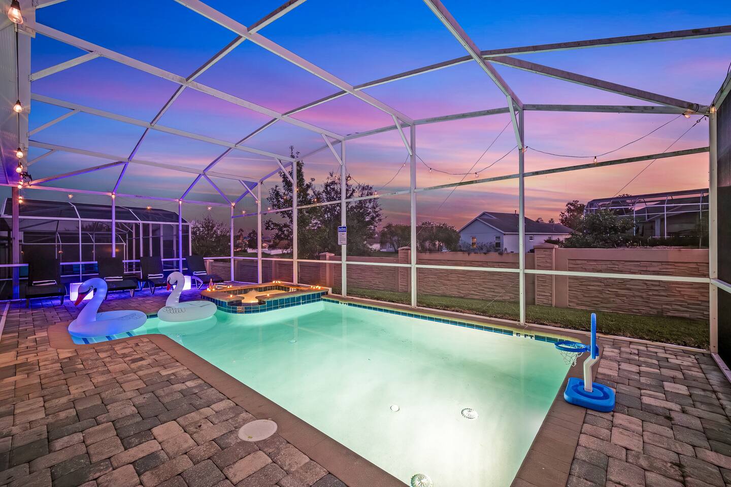 Lit pool with basketball hoop and spa inside the screened enclosure at dusk