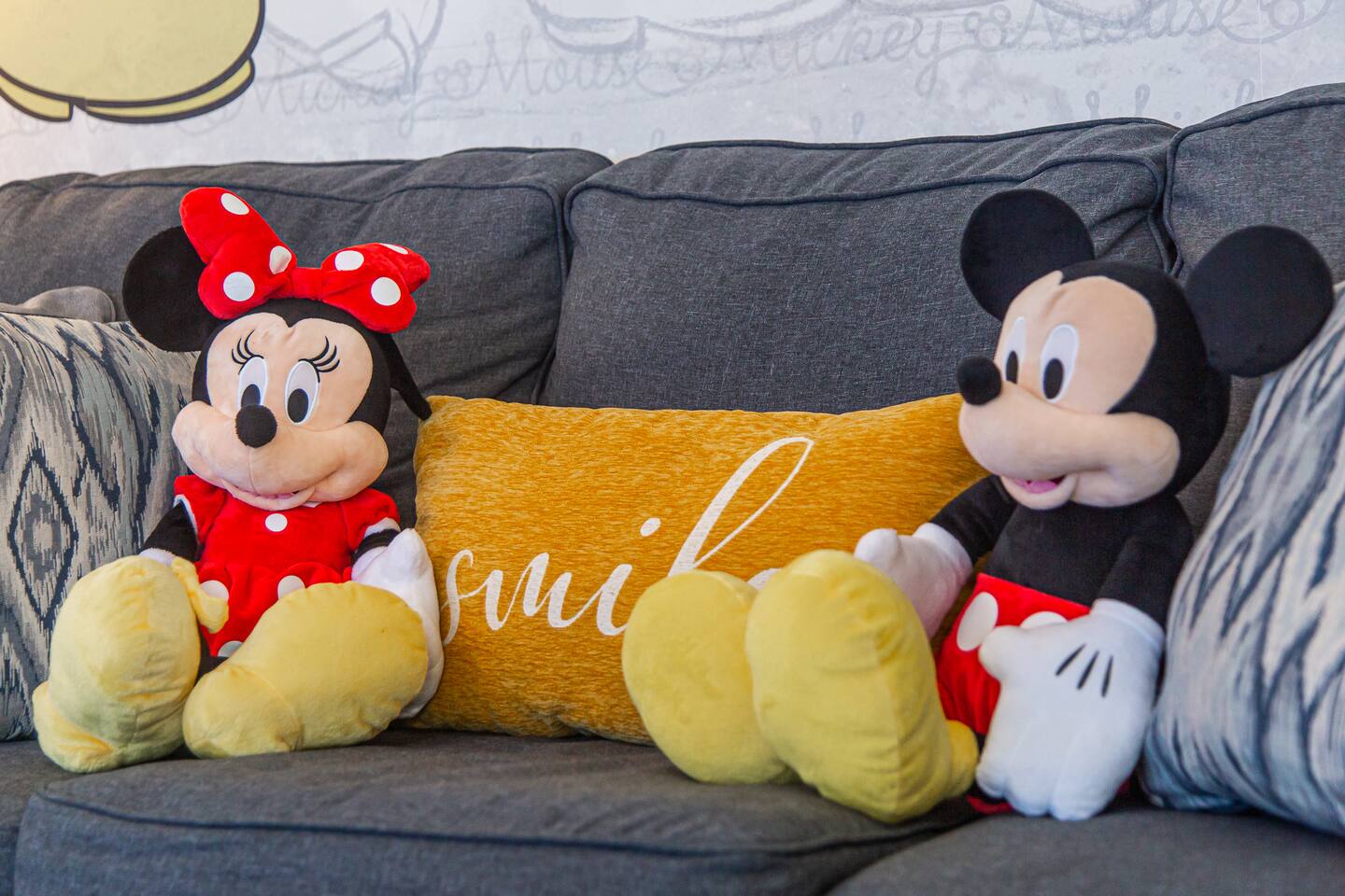 Minnie and Mickey Mouse plush toys flanking a smile pillow on a gray sofa.