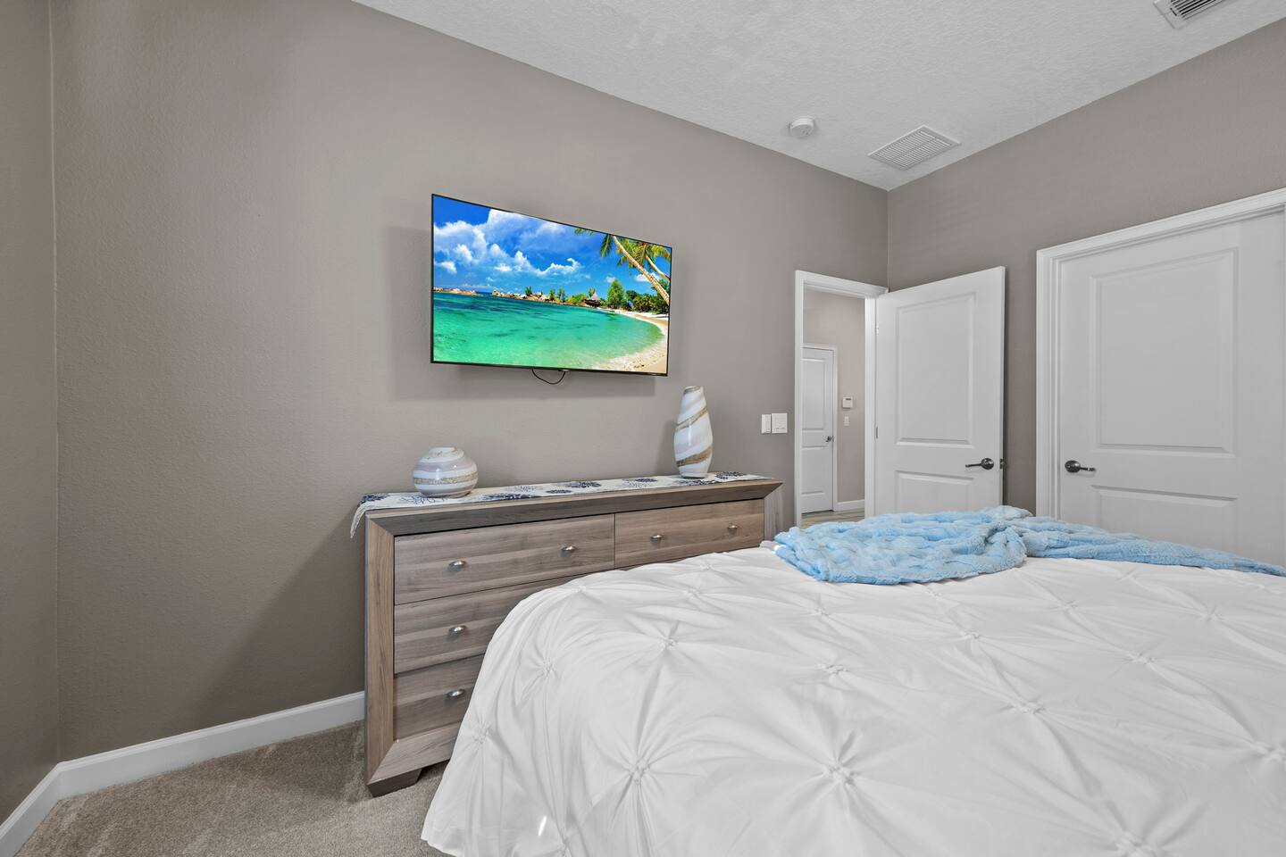 Same bedroom showing the dresser, wall-mounted TV, and doors to the ensuite bath