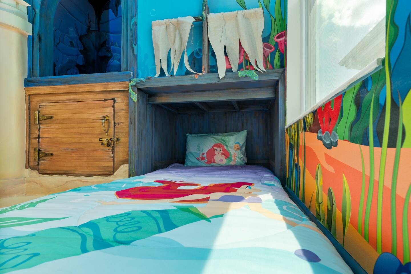 Twin sleeping nook with mermaid bedding beside a latched wooden hatch door in th