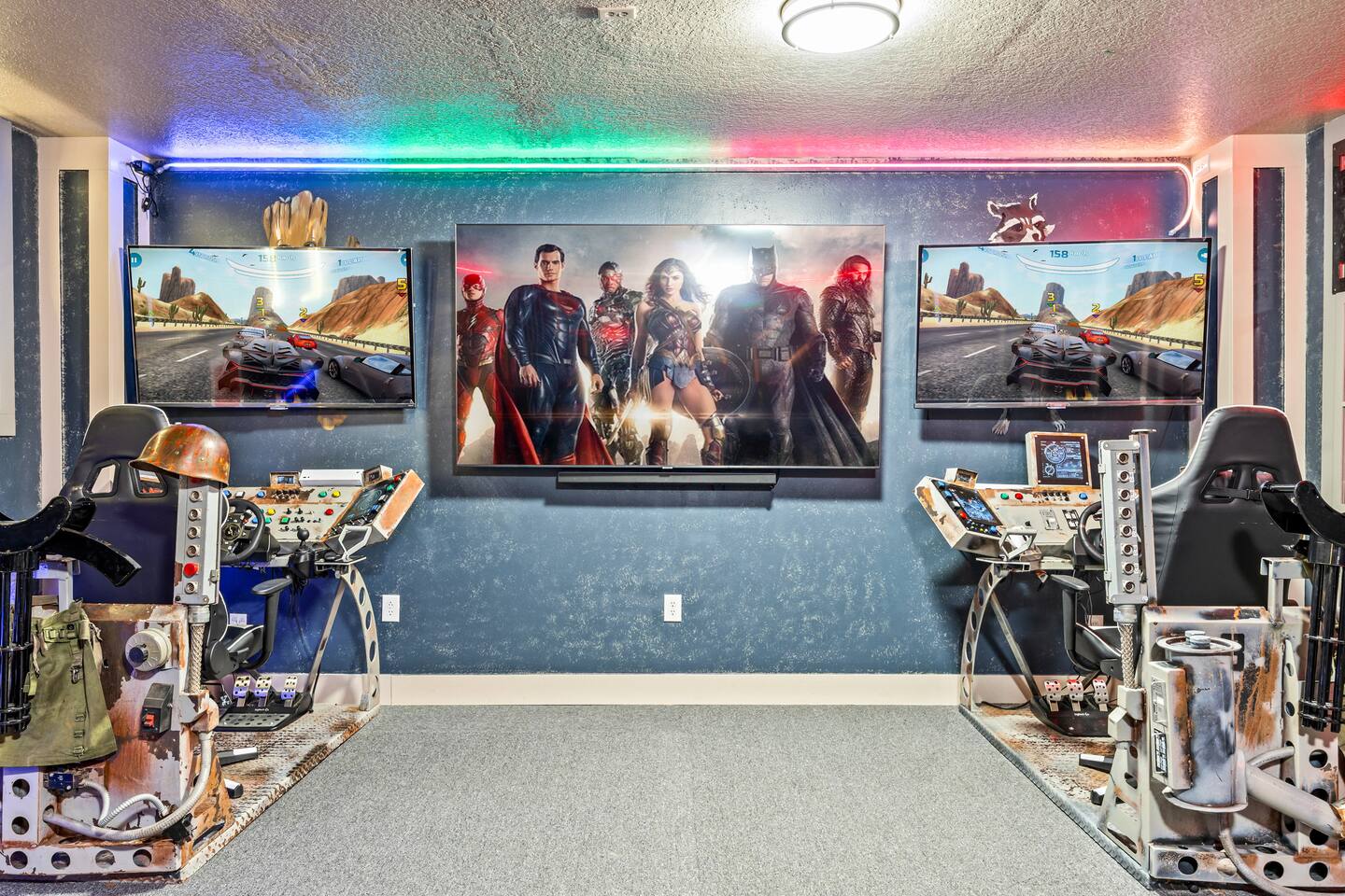 Game room with two racing simulators flanking a large TV and a superhero mural.